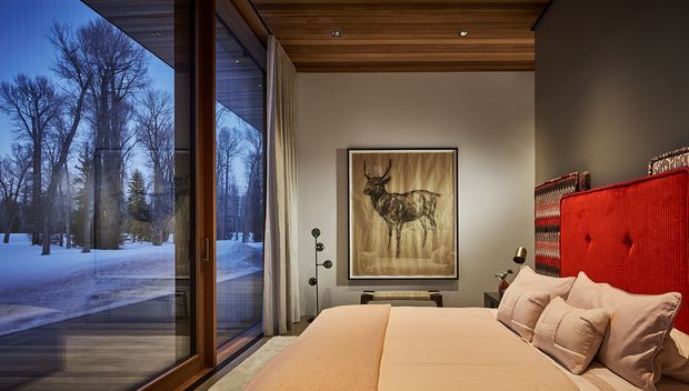 A warmly lit bedroom with a large bed dressed in soft beige bedding, a vivid red upholstered headboard, and a framed deer artwork. Full-height glazing and a timber-framed sliding door reveal a snow-covered wooded landscape outside. The room features a rich timber slatted ceiling, neutral walls, layered curtains, and a compact sculptural floor lamp, creating a refined alpine retreat atmosphere.