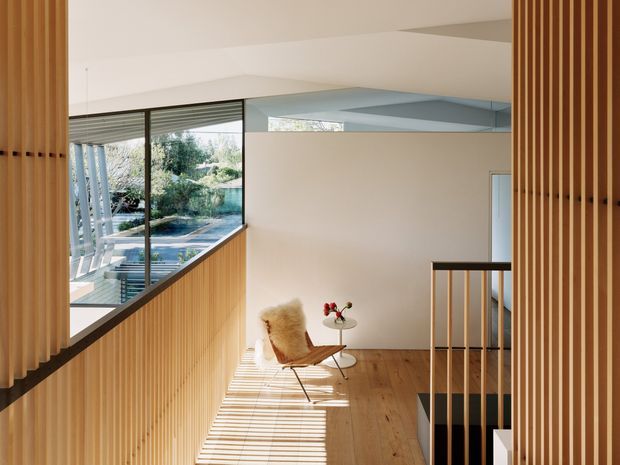 A bright, double-height modern interior landing or hallway with warm vertical timber slat screens and balustrades, pale plaster walls, timber flooring, and extensive glazing overlooking greenery. A sculptural lounge chair and small side table sit in the sunlit circulation space, while strong linear shadows from the slats fall across the floor. A stair opening with timber balustrade is visible at right.