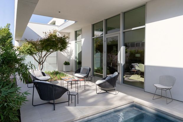 A bright contemporary courtyard terrace beside a small swimming pool, featuring white rendered walls, broad overhanging roof planes, tall glazed doors and windows, and minimalist outdoor furniture arranged around low coffee tables. Landscaped planting softens the courtyard edges, while an interior dining area is visible through the glazing.