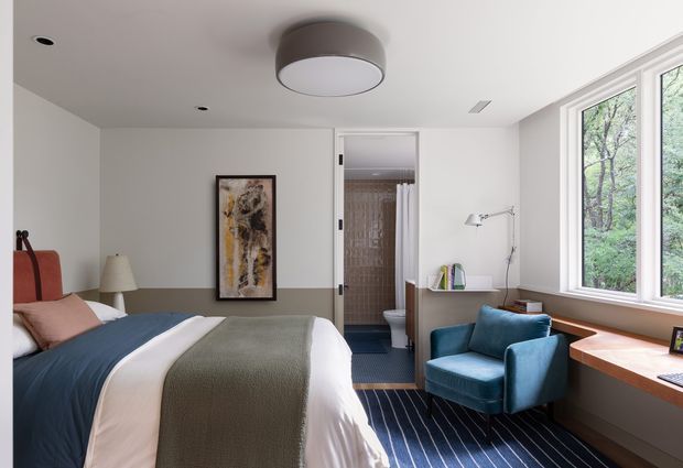 Bright contemporary bedroom with a large bed dressed in white, blush, blue and olive-green bedding, a teal upholstered armchair, and a built-in timber desk beneath expansive windows. A compact ensuite bathroom is directly visible through the open doorway, while abstract wall art, recessed ceiling lights and a contemporary flush-mount ceiling fixture complete the room.