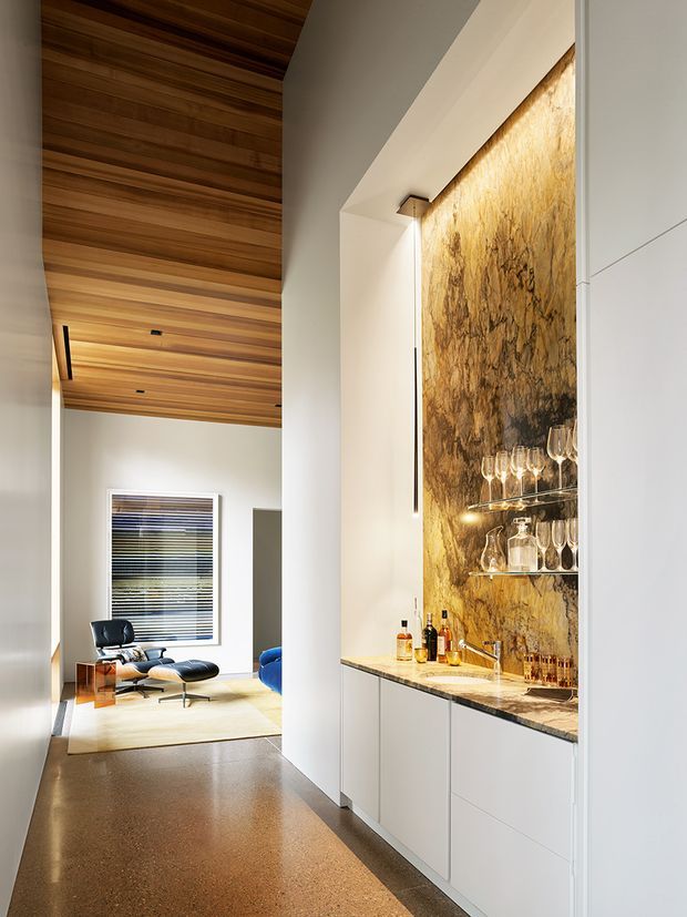 A refined open-plan interior featuring a built-in home bar along a hallway or living area. The bar has sleek white cabinetry, a dark stone benchtop, a dramatic backlit amber-toned stone feature wall, glass shelving, and a collection of glassware and bottles. Adjacent is a comfortable lounge with a black leather chair and ottoman beneath a striking warm timber-lined ceiling. The space has polished warm-toned flooring, white walls, and clean contemporary detailing.