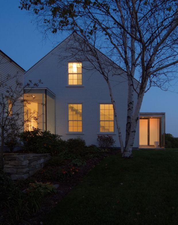 Twilight exterior view of a gabled house clad in pale horizontal weatherboards. Warm light glows through divided-pane windows and a recessed entrance, with a small glazed flat-roofed extension visible on the right. A mature birch tree, lawn, planted garden beds, and a low rough-stone retaining wall frame the facade.