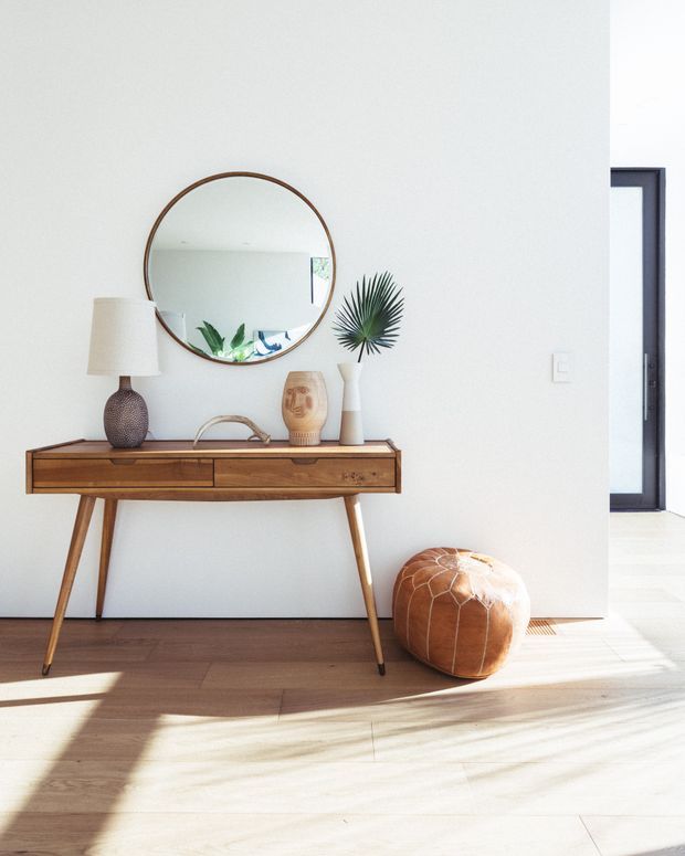 Bright, minimalist entryway with a mid-century-style wooden console table against a white wall. The console is styled with a round wood-framed mirror, table lamp, ceramic vessels, and greenery, with a tan leather pouf on the timber floor. A dark-framed doorway and strong natural sunlight are visible at right.