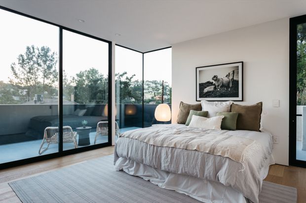 Bright contemporary bedroom with a large bed dressed in layered white and beige linens, framed black-and-white artwork, and a minimalist bedside lamp. Floor-to-ceiling black-framed glazing wraps around the room and opens onto a small balcony or terrace with outdoor seating and views of trees. The space has pale timber flooring, white walls, and a soft neutral rug.