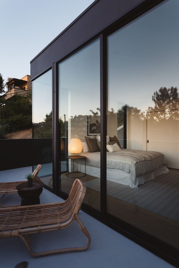 A modern bedroom viewed from a compact outdoor terrace through full-height dark-framed glazing. The room contains a low platform bed with layered white bedding, neutral pillows, a glowing spherical bedside lamp, and a small round side table. The terrace has woven lounge chairs, a dark planter, and smooth blue-grey paving. Reflections and surrounding trees are visible in the glass, while the architecture is defined by crisp geometric forms and expansive glazing.