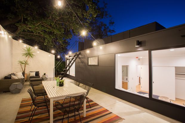 A contemporary outdoor courtyard or terrace at dusk, furnished with a dining table, woven dining chairs, lounge seating, and a colourful striped rug. Festoon lighting hangs among mature trees, while a dark-clad modern building with large illuminated windows and an exterior stair forms the backdrop. The space feels intimate, warm, and connected to the interior.