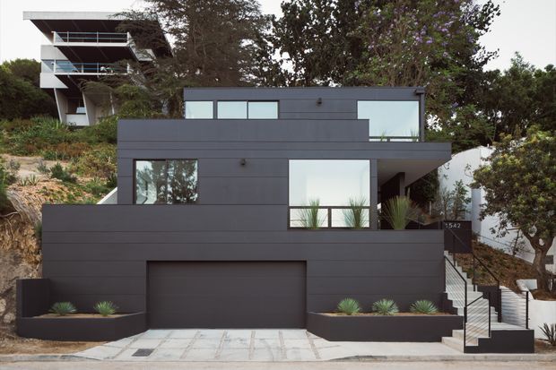 Street-facing view of a contemporary multi-level residence on a sloped, landscaped site. The main house has a dark charcoal horizontal-cladding facade, broad rectangular glazing, an integrated garage, stepped entry stairs, and simple low-maintenance planter beds. A taller neighbouring or upper dwelling is visible behind it.