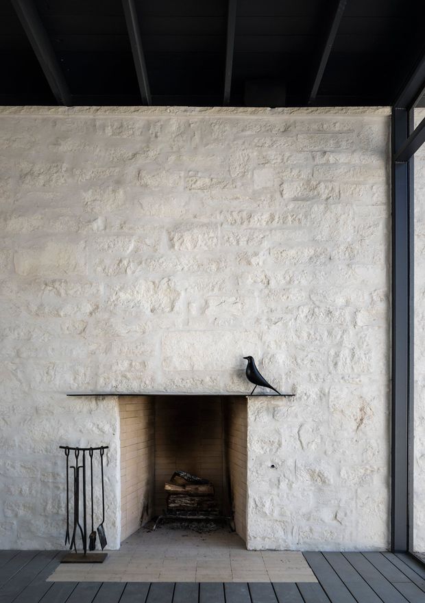 A minimalist interior fireplace set into a rough white-painted masonry wall. The deep open hearth has pale brick lining and a simple dark mantel shelf with a black bird sculpture. A black exposed-beam ceiling and dark timber flooring frame the composition, while full-height glazing or a dark-framed opening appears at the right.