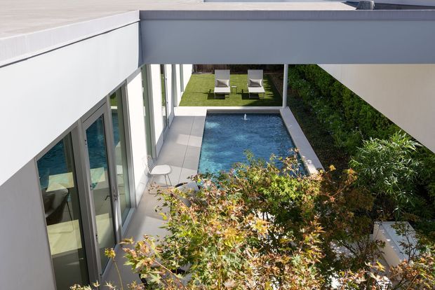 View into a modern, enclosed courtyard framed by clean white and grey architectural volumes. The space contains a narrow rectangular swimming pool, paved surrounds, lawn with loungers, dense planted screening and full-height glazed doors. The image is taken from an elevated position, emphasizing the geometric courtyard layout and strong indoor-outdoor connection.