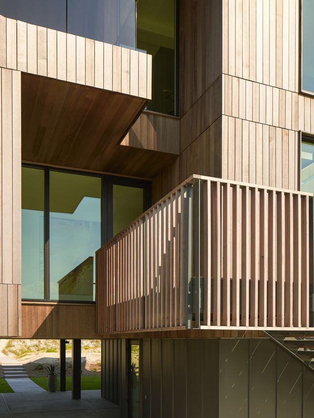 Close-up exterior view of a contemporary multi-level residential building with extensive vertical timber cladding, dark metal or painted panels, large glazed openings, projecting timber soffits, and a raised balcony enclosed by a vertical timber-slat balustrade. The image emphasizes the facade, balcony, and cantilevered architectural forms.