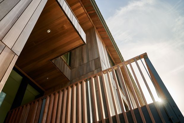 Low-angle exterior view of a contemporary multi-level building featuring extensive vertical timber cladding, a warm timber soffit and balcony decking, and a slatted timber-and-metal balustrade. The image emphasizes the projecting balcony, layered facade, and pitched roof edge against the sky.