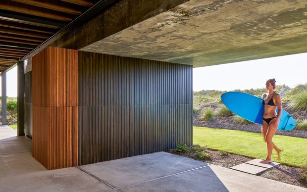 A covered outdoor passage beside a modern building, featuring a projecting concrete slab ceiling, dark vertically ribbed cladding, warm timber screening, concrete paving, and landscaped coastal planting. A person carrying a surfboard is visible in the garden beyond.