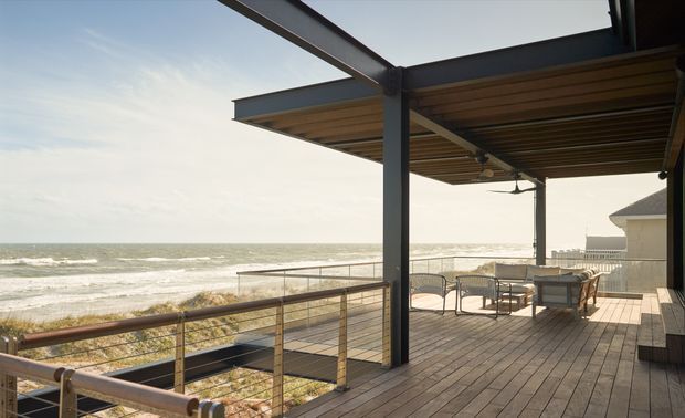 A spacious elevated timber deck overlooking the ocean, with a dark steel-framed covered pergola, exposed timber soffit, outdoor seating and a balustrade. The open-sided terrace has direct beachfront views and a relaxed coastal setting.