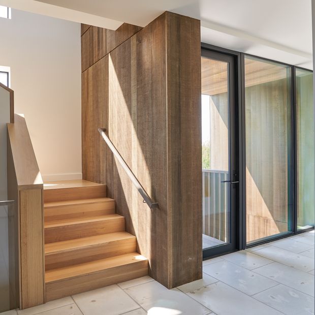 Bright contemporary interior stairway with warm vertical timber wall panelling and matching timber stair treads. A simple metal handrail runs along the paneled wall, while large black-framed glazed doors and windows open to an adjoining outdoor area. Pale large-format tiled flooring, white walls and strong natural sunlight create a clean, warm, minimal composition.