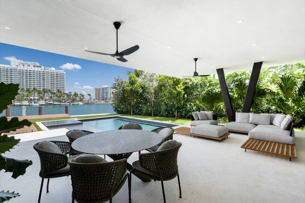 A spacious covered outdoor entertaining area overlooking a swimming pool and waterfront. The space features a round dining table with woven chairs, a pair of light upholstered lounge sofas, a slatted timber coffee table, concrete-look flooring, ceiling fans, recessed lighting, and dark angled structural posts. Tropical planting surrounds the pool, with high-rise buildings visible across the water.