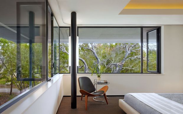 Bright modern bedroom with expansive black-framed corner glazing overlooking trees. The room contains a low upholstered bed, a mid-century-style lounge chair and small side table, white walls, dark flooring, and a recessed ceiling feature with a warm yellow accent. The glazing and reflected surfaces create a strong indoor–outdoor connection.