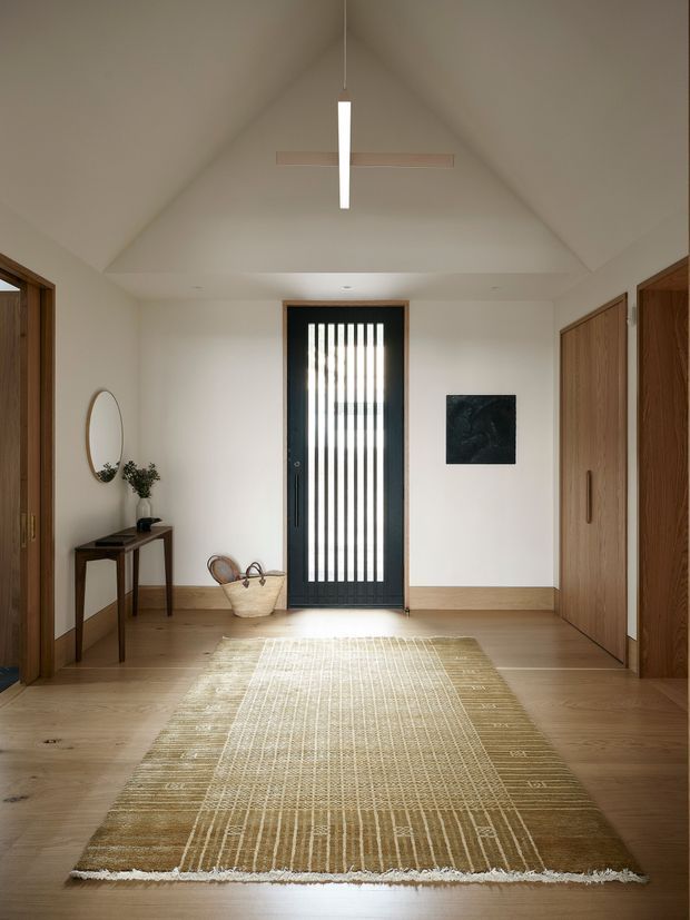 A warm, minimalist residential entry hall with a high vaulted ceiling and a central black-framed glazed door featuring vertical privacy slats. Natural timber doors, built-in cabinetry, and timber flooring surround a woven neutral rug. A slim console table, round mirror, small plant, basket, and understated wall artwork add restrained decoration. A linear pendant hangs beneath the vaulted ceiling, creating a calm and welcoming entry.