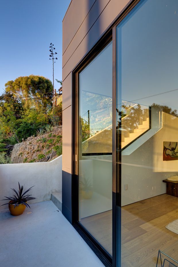 Vertical architectural photograph showing the corner of a contemporary home where a large black-framed glazed opening connects an outdoor concrete terrace with a bright interior. The facade uses smooth horizontal panel cladding, while the interior reveals warm timber flooring, a staircase, white walls, and built-in joinery. A planted garden and low rendered boundary wall sit beside the terrace, with reflections visible in the glazing.