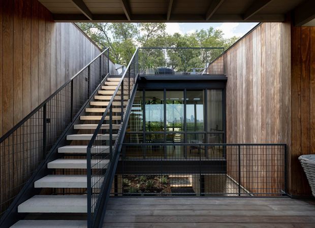 A contemporary timber-lined courtyard or terrace viewed from an elevated deck, featuring an open-riser exterior staircase with black metal railings, dark-framed glazed doors, timber cladding, and a partially covered overhead structure. The space appears to connect multiple levels around a planted lower courtyard.