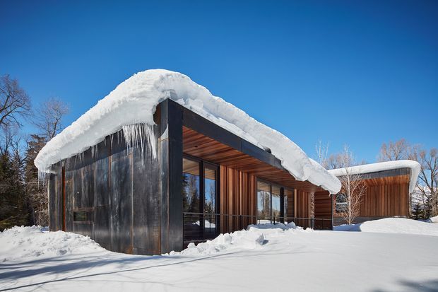 A low, modern house set in a snowy wooded landscape. The building has dark vertical cladding, extensive floor-to-ceiling glazing, warm timber soffits and recessed outdoor living areas. Heavy snow blankets the flat or gently curved roof forms, with long icicles along the eaves. Bright blue sky and bare trees emphasize the alpine winter setting.
