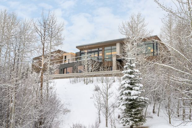 A contemporary multi-level house set on a steep, snow-covered wooded slope. The building features broad floor-to-ceiling glazing, dark metal-framed windows and balustrades, warm timber-toned wall sections, and projecting flat roof planes. Bare deciduous trees and snow-covered evergreens surround the elevated residence.