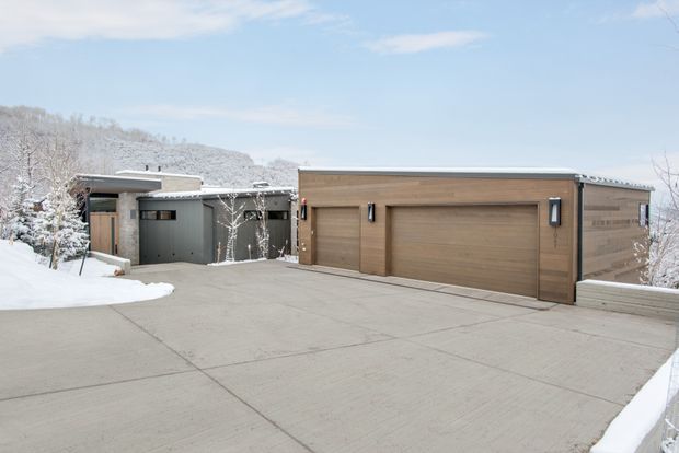 Snow-covered exterior view of a contemporary flat-roof home and attached multi-car garage. The broad concrete driveway dominates the foreground, with warm wood-toned garage doors, dark grey wall cladding, minimal rectangular forms, and snowy trees on a hillside in the background.