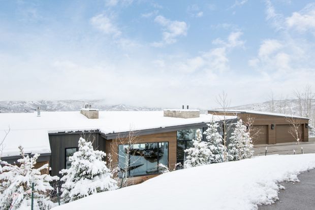 Exterior view of a low-slung contemporary house set in a snowy, mountainous landscape. The building has a broad flat roof, dark and warm brown horizontal cladding, extensive glazing, and a partially recessed entry or living area. Snow-covered conifers and a paved driveway frame the foreground.