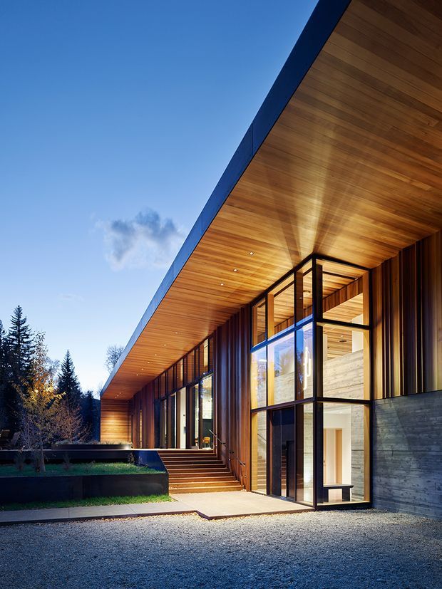 Exterior architectural photograph of a contemporary single-storey residence with a dramatic deep pitched roof and broad timber-lined soffit. Floor-to-ceiling glazing, dark vertical timber cladding, exposed concrete wall elements, and a recessed entry define the facade. Landscaped raised garden beds and a gravel forecourt sit beside the building, with trees and blue sky in the background.