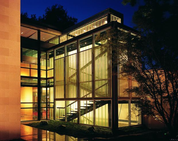 Night-time exterior view of a contemporary multi-storey building with extensive floor-to-ceiling glazing, dark structural framing, illuminated interior circulation and staircases, and a warm stone-clad wall at left. The glazed central volume creates a prominent vertical atrium-like composition, with surrounding trees and reflective paving visible at ground level.