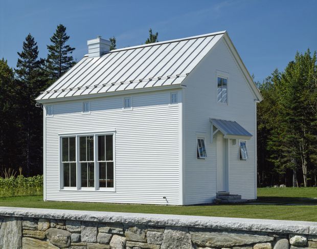 Exterior view of a compact white weatherboard house with a steep standing-seam metal gable roof, small covered entry, chimney, multi-pane windows, and a low stone retaining wall in front. The house sits in a wooded, grassy setting under clear daylight.