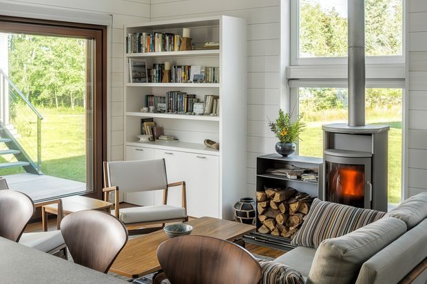 Bright, cozy living room with white horizontal wall panelling, extensive built-in white bookshelves and cupboards, large windows and glazed doors overlooking greenery, a freestanding wood-burning stove with log storage, a timber coffee table, lounge seating, and mid-century-inspired chairs. The room has abundant natural light and a warm contemporary cabin-like character.