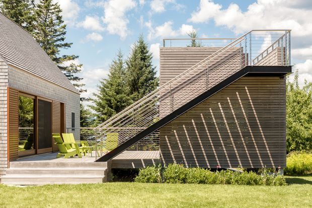 Exterior view of a contemporary residential property with a steeply sloped outdoor stair leading to a rooftop terrace or upper deck. The building combines horizontal timber cladding, dark structural elements, and metal cable-style railings. A shingle-clad adjacent volume has large glazed doors opening onto a timber deck with bright outdoor chairs. The setting includes lawn, established trees, and a bright partly cloudy sky.