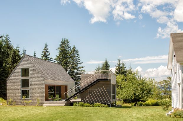 Exterior view of a contemporary shingle-clad residential property in a landscaped grassy setting. The main building has a steeply pitched gable roof, tall vertical glazing, a compact deck and an exterior stair leading to an elevated terrace. Mature evergreen trees and a partly visible neighbouring wing frame the composition under a bright blue sky.