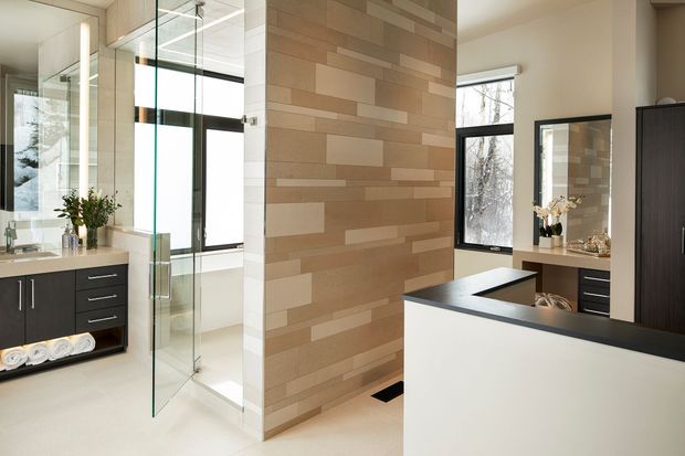 Contemporary bathroom with a freestanding bathtub behind a clear glass shower enclosure, a feature wall clad in horizontally arranged beige and taupe rectangular tiles, dark wood vanity cabinetry, pale flooring, and large black-framed windows. A mirrored vanity area and built-in storage are also visible.