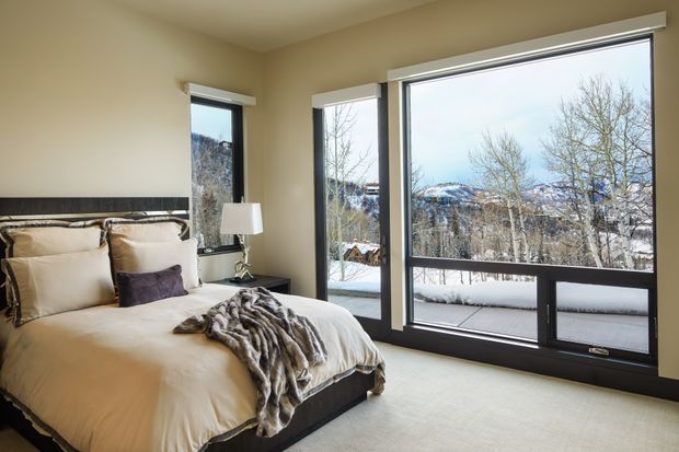 A warm, modern bedroom with a large upholstered bed, layered neutral bedding, dark wood headboard, bedside lamp, and floor-to-ceiling corner glazing. Snow-covered trees and mountains are visible outside, with access to a balcony or terrace.