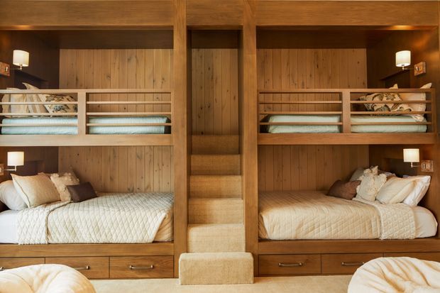 A symmetrical, built-in bunk bedroom with two lower double beds, upper sleeping bunks, integrated shelving, drawers, and a central timber staircase. Warm wood panelling and joinery dominate the room, complemented by cream quilted bedding, neutral cushions, and small bedside lamps. The compact, highly crafted design has a cozy, refined cabin-like atmosphere.