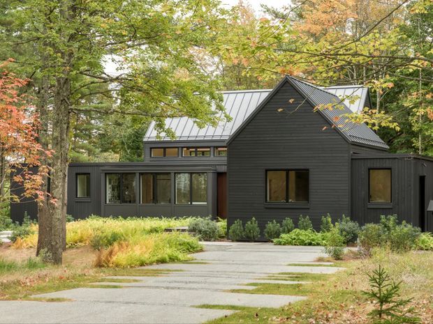 A contemporary single-storey dark-clad house set within a mature wooded landscape. The building has a prominent pitched gable volume, standing-seam metal roofing, horizontal dark siding, large glazed openings, and a connected lower wing. A paved approach passes through landscaped planting and autumnal trees.