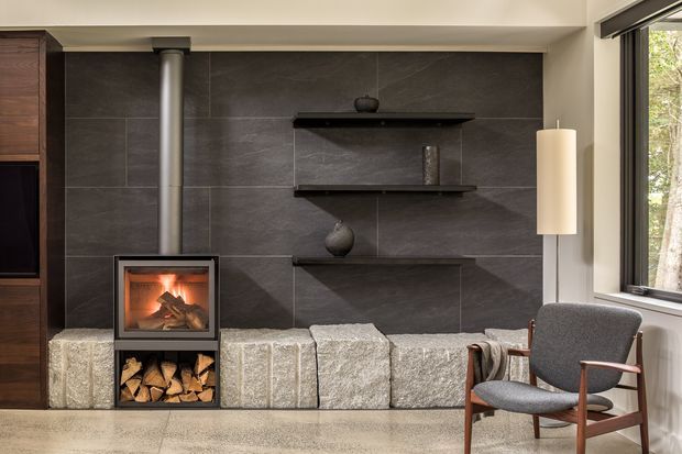 A refined modern living room centered on a built-in fireplace with a wood-storage compartment below. The fireplace is set against a dark large-format tiled feature wall with floating dark shelves and sculptural decor, while pale rough-cut stone blocks form a low hearth or bench. Full-height glazing at the right opens the room visually to trees outside. A timber-framed upholstered lounge chair, pendant light, and dark timber cabinetry complete the warm, contemporary composition.