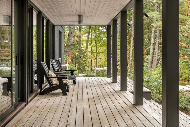 A covered timber deck running alongside a modern glazed dwelling, with dark structural posts, full-height sliding glass doors, a timber-lined ceiling, and black lounge chairs. The deck overlooks a wooded landscape with mature trees and dense planting.