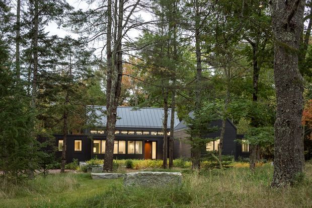 A low, contemporary dark-clad house nestled among mature trees and natural grasses. The building has a simple pitched metal roof, expansive warm-lit windows, and a modest landscaped foreground with large natural stone elements. The wooded setting dominates the scene, creating a secluded cabin-like residential exterior.