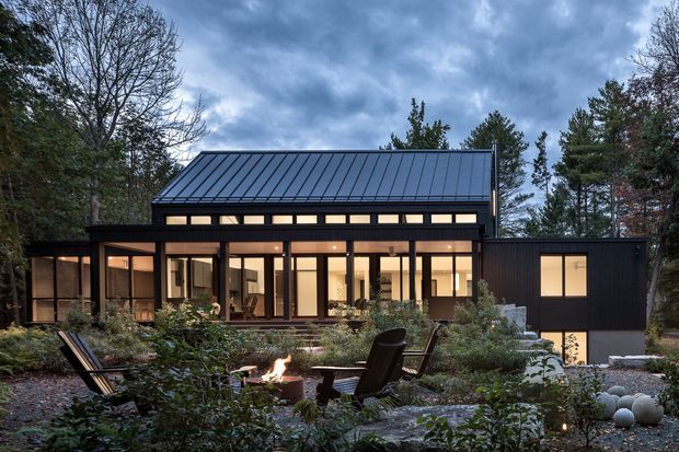 A modern dark-clad house set within a landscaped, wooded garden at dusk. The low-slung building has extensive floor-to-ceiling glazing, a prominent standing-seam metal gable roof, warm interior lighting, and a projecting glazed living area. In the foreground, a landscaped outdoor seating area surrounds a small fire pit.