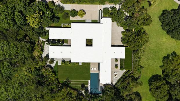 Aerial top-down view of a large modern residence set within landscaped grounds. The building has a strongly geometric, cross-like plan with broad flat white roof planes, a central square roof opening, paved paths and terraces, and a narrow blue swimming pool extending from one side. Mature trees and expansive lawns surround the property.