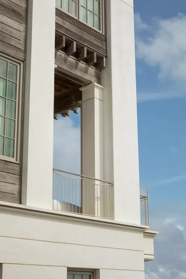 Close-up exterior view of a refined multi-storey building facade featuring white rendered columns and walls, weathered timber cladding, tall multi-pane windows, decorative timber brackets, and a slender metal balcony balustrade. Blue sky is visible through the open architectural recess.