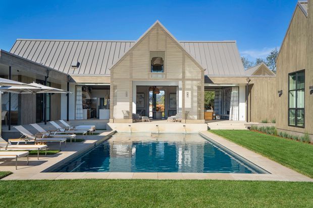 A contemporary residential courtyard centered on a rectangular swimming pool, with a light-toned gabled and metal-roofed house surrounding the pool on three sides. The facade combines pale horizontal cladding, large glazed openings, and dark-framed windows. Poolside loungers and umbrellas occupy the paved terrace, while a neatly maintained lawn borders the foreground.