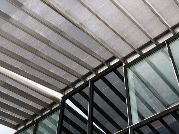 Close-up architectural view of a translucent covered roof or canopy with repeated structural beams, meeting a dark-framed glazed facade. The image emphasizes exposed structural members, linear geometry, filtered daylight, and reflective glass; no identifiable room interior is visible.