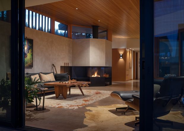 A refined open-plan living room viewed through dark-framed glazing, featuring a dramatic central fireplace with a black surround, warm timber-lined ceiling, textured plaster walls, expansive high-level windows, a neutral sectional sofa, mid-century-style lounge chair and ottoman, a sculptural timber coffee table, patterned rug, and a cowhide-style floor covering. The room has warm ambient lighting and a sophisticated, cozy atmosphere.