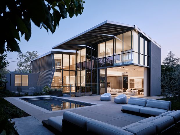 A dusk exterior view of a large contemporary multi-level residence with extensive floor-to-ceiling glazing, a strong exposed steel structural grid, and a projecting flat roof with deep overhangs. The house opens onto a paved terrace with a swimming pool and substantial outdoor lounge seating. Warm interior lighting contrasts with the cool blue-grey exterior setting, while mature planting frames the property.