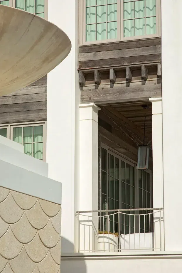 Close-up exterior architectural detail of a light-coloured multi-storey facade. The composition features weathered horizontal timber cladding, tall divided-light windows with decorative wavy curtains, white rendered or painted columns, a timber-lined recessed balcony or entry, projecting timber brackets, and a delicate metal balustrade. A curved pale architectural element and scalloped wall detailing appear in the foreground. The image is brightly lit with a refined coastal or traditional character.