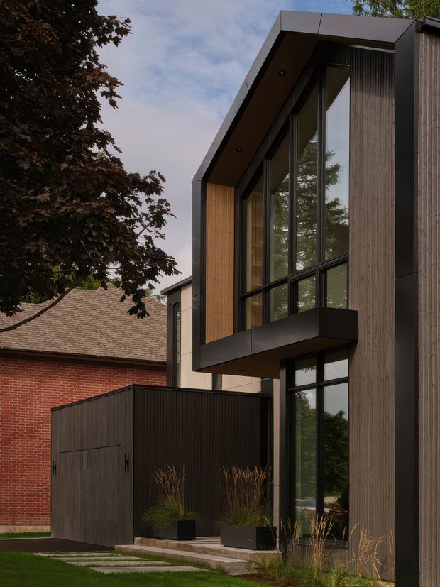 Exterior view of a contemporary multi-level house with a sharply angled roofline, floor-to-ceiling black-framed glazing, vertical timber-textured wall cladding, and dark metal trim. A compact dark-clad outbuilding or garage sits in front, with landscaped grasses and paving. A red-brick traditional house is visible in the background.