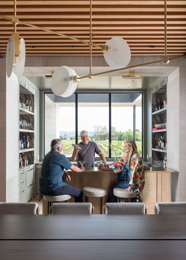 A refined home bar or entertaining area viewed from a dining table, featuring a central island with stools, extensive open bottle and glass shelving on both sides, a large picture window, warm timber slatted ceiling, and sculptural brass-and-white pendant lighting. Three people are gathered casually at the bar. The space has a contemporary, warm, hospitality-inspired design with dark-framed glazing and muted neutral finishes.